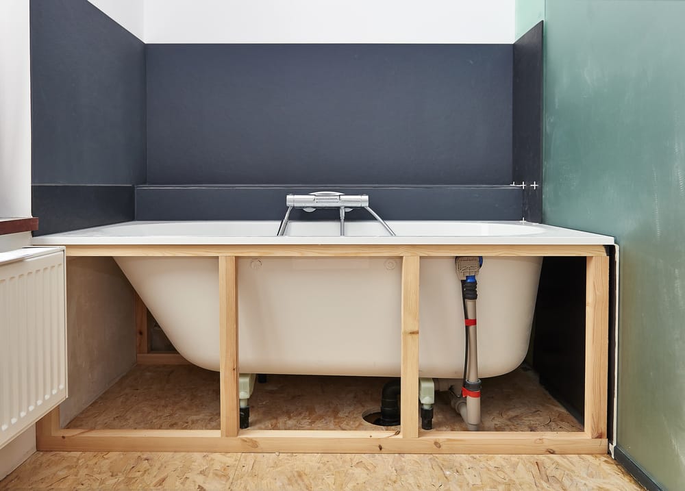 A partially installed bathtub with exposed plumbing, framed by an unfinished wooden support structure. The surrounding walls are a mix of dark and light colors.