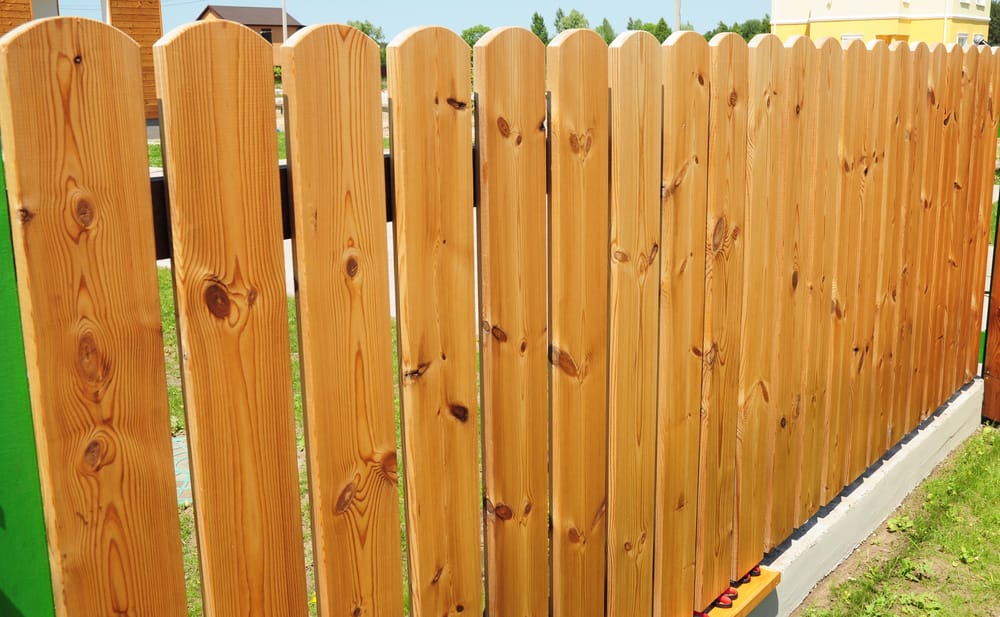 A wooden fence made of vertical planks with a natural finish stands outdoors, separating a grassy area from neighboring properties.