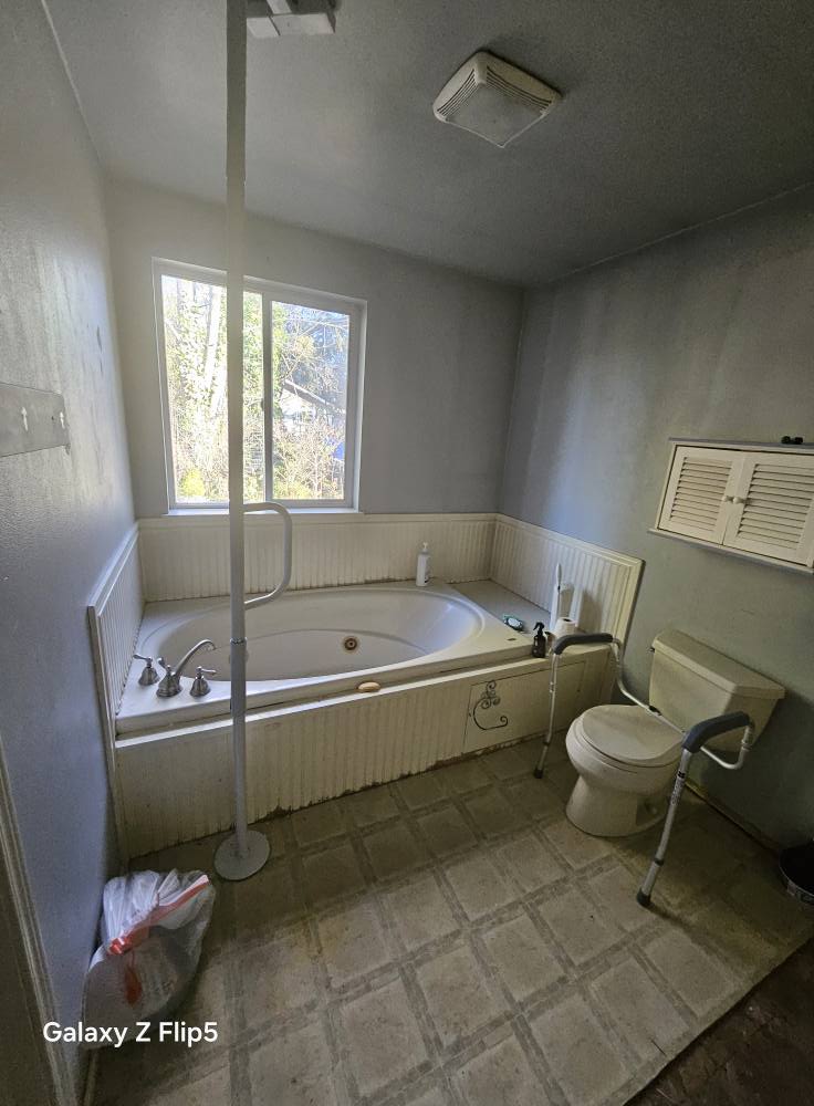 A bathroom with a large tub, nearby window, toilet with support rail, and a waste bin. Walls and ceiling are painted gray. A light fixture is on the ceiling.