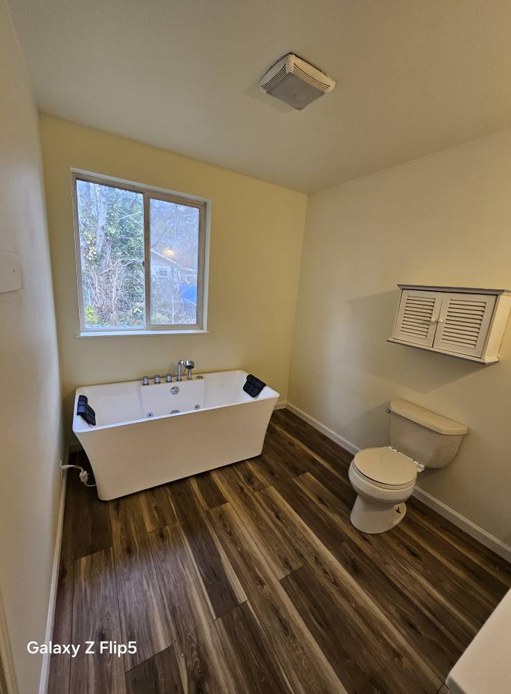 Bathroom with freestanding bathtub, toilet, and wall cabinet. Wood-like flooring and a window overlooking trees.