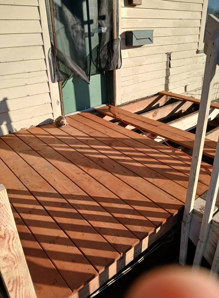 A wooden deck under construction with a partially assembled railing. A door and a window with a torn screen are visible on the adjacent building wall.