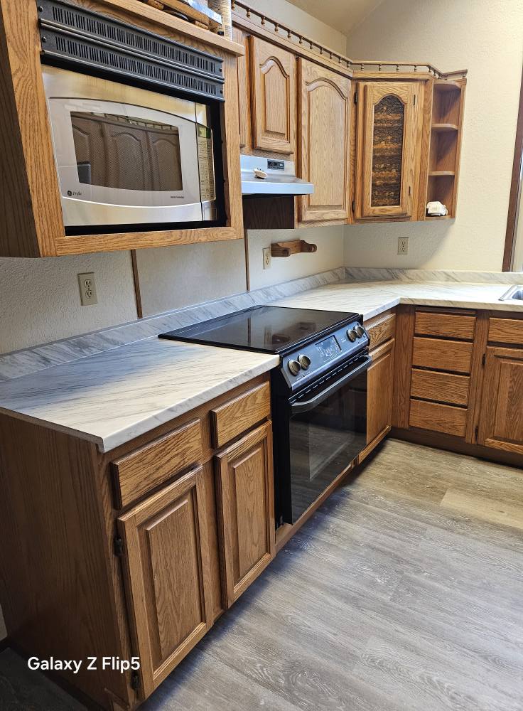 A kitchen with wooden cabinets, a black electric stove, and a wall-mounted microwave above the countertop. The floor is gray laminate.