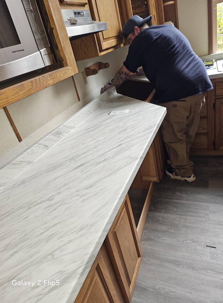 A person installs a marble countertop in a kitchen, with wood cabinets and a microwave visible.
