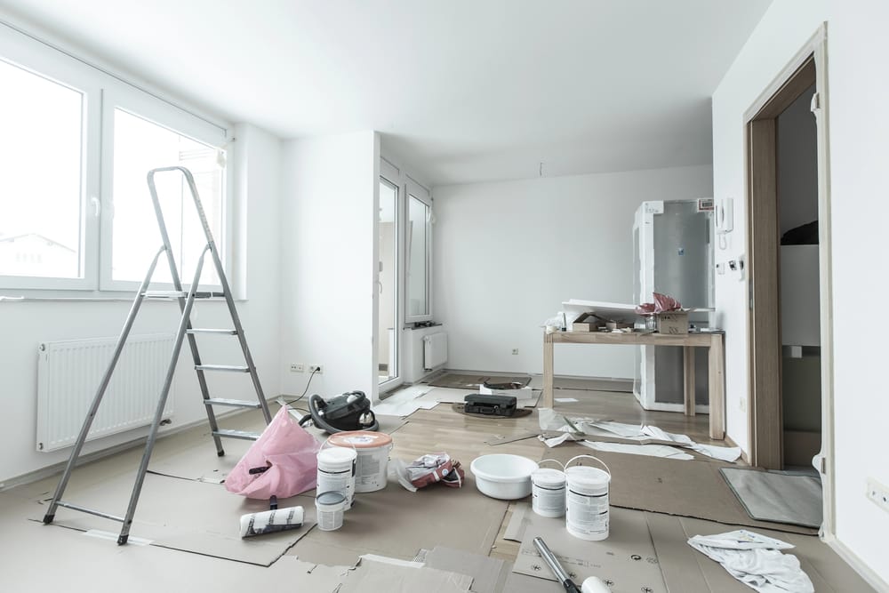 Room under renovation with a ladder, scattered paint buckets, tools, and cardboard on the floor. A table holds supplies, and there's a partially visible window and door.