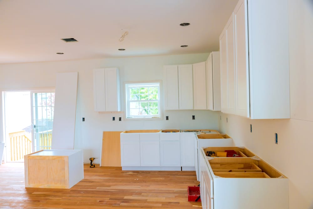 A kitchen under construction with white cabinets partially installed and wooden flooring. The room is well-lit by natural light from a window and a glass door.