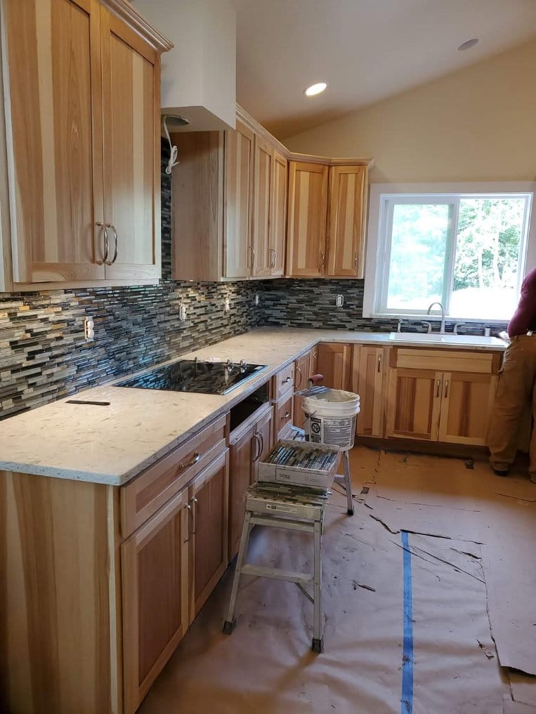 Kitchen under renovation with wooden cabinets, stone backsplash, and countertops being installed; tools, materials, and a step ladder hint at ongoing kitchen remodeling. A person stands near the window overseeing the progress.